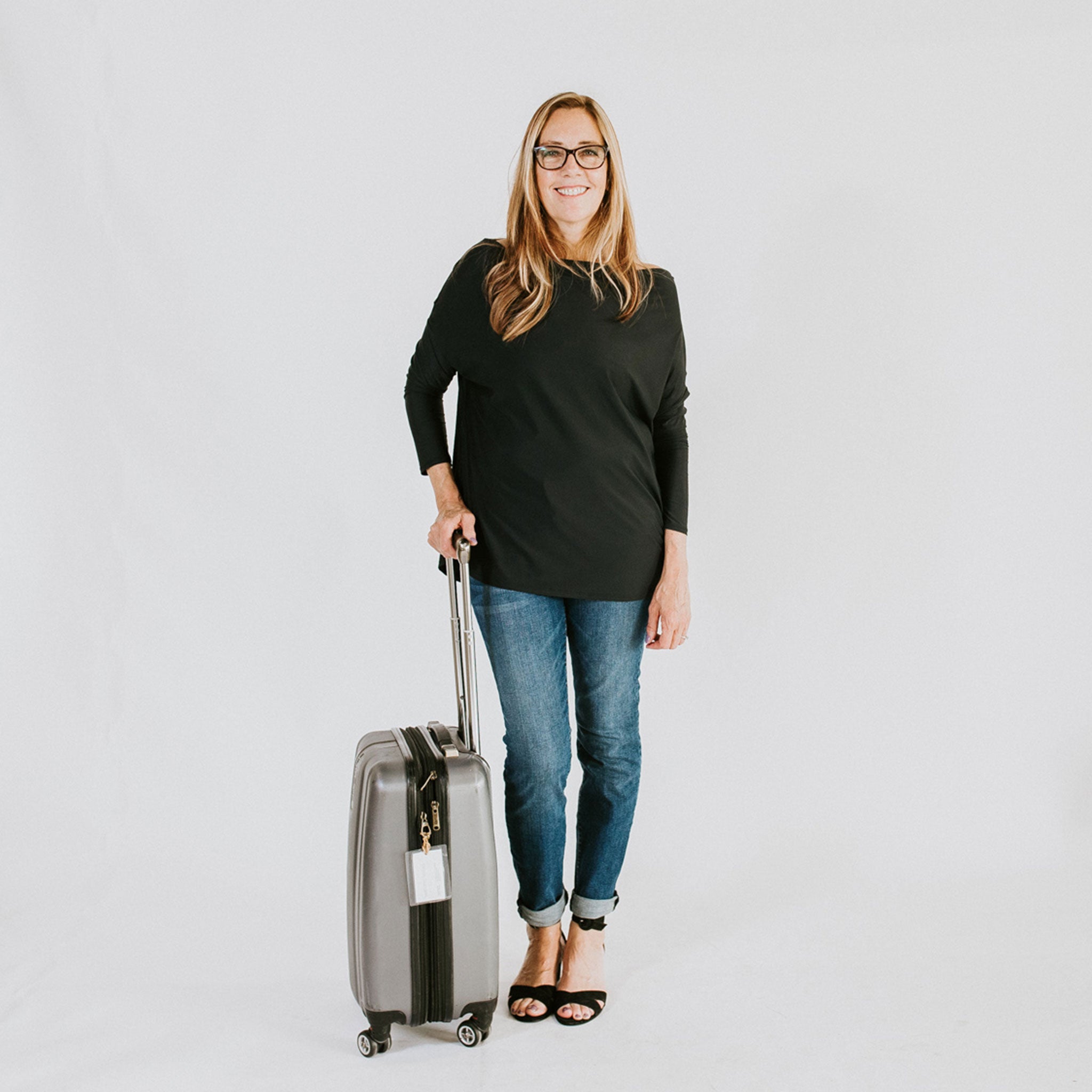 happy woman wearing a black long sleeve shirt and jeans while resting their hand on their rollable luggage handle 