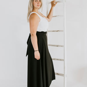 side profile of a woman wearing a white tank top and a black floor length wrap skirt while resting her arm on a ladder