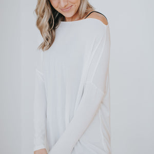 smiling woman wearing white long sleeve shirt draped off one shoulder, the fabric is thin and you can see her black tank underneath